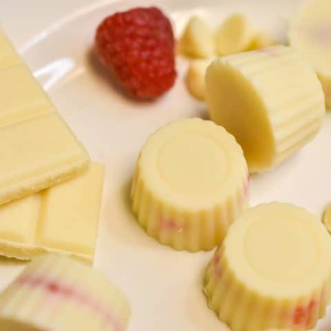 white chocolate raspberry fat bombs on a white plate with raspberries and chocolate bars