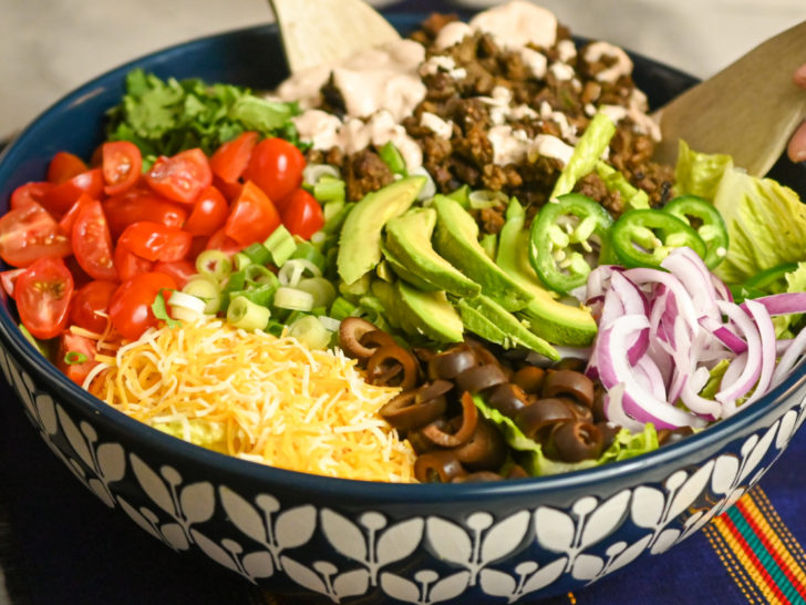keto ground beef taco salad being tossed in a large blue and white salad bowl with wooden utensils