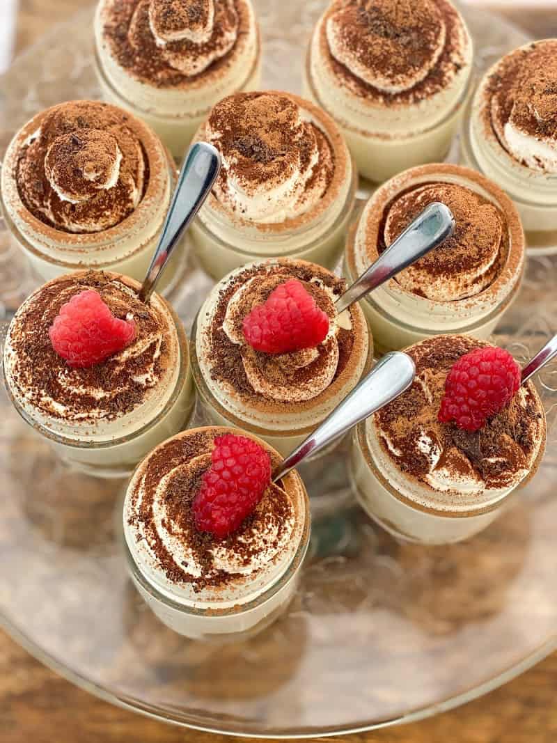 individual keto tiramisu cheesecakes with spoons and raspberries on a glass stand on top of a wooden board