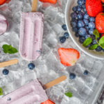 keto berries and cream popsicles on ice with popsicle mold and bowl of berries on the right