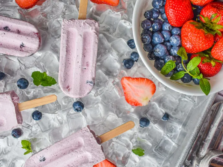 keto berries and cream popsicles on ice with popsicle mold and bowl of berries on the right