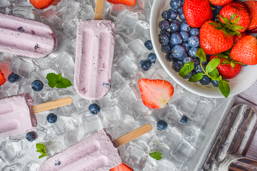 keto berries and cream popsicles on ice with popsicle mold and bowl of berries on the right