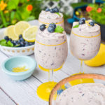 keto blueberry cheesecake mousse in glass cups topped with blueberries with a bowl of blueberries and lemon and plants in the background