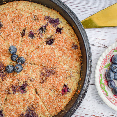 keto blueberry toasted coconut skillet cake in cast iron skillet with dish of blueberries and cake server on the right