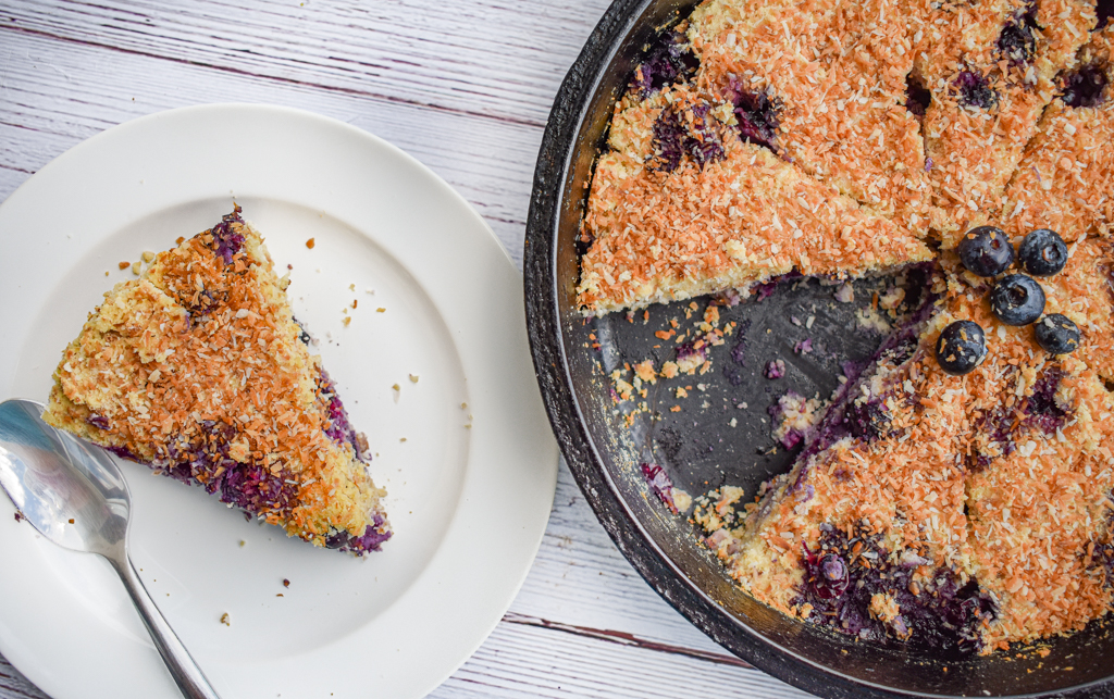 keto blueberry toasted coconut skillet cake in cast iron skillet with one slice missing and slice on white plate on the left