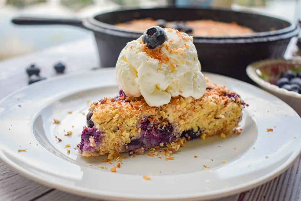 keto blueberry toasted coconut skillet cake slice topped with ice cream with cast iron and blueberries in the background