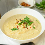 keto roasted cauliflower soup in white bowl on white plate with black spoon and garnish in background