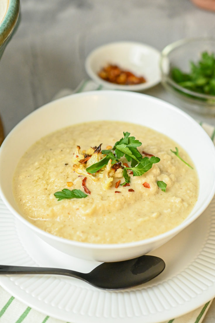 keto roasted cauliflower soup in white bowl on white plate with black spoon and garnish in background