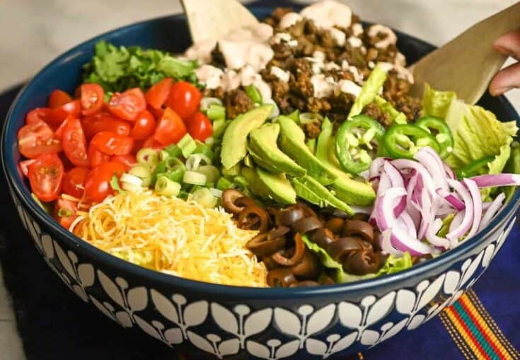 keto ground beef taco salad being tossed in a large blue and white salad bowl with wooden utensils